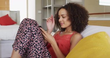 Afro-Amerikan kadın, akıllı telefon tebessümü kullanarak yatakta oturuyor. Karantina süresince evde tecrit altında kalmak.