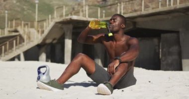 Yorgun Afrikalı Amerikalı adam sahilde oturuyor, şişeden içiyor, dışarıda egzersiz yapıyor. fitness, sağlıklı ve aktif yaşam tarzı konsepti.