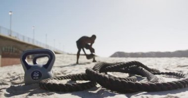 Afrika kökenli Amerikalı adam plajda egzersiz yaparken topu kaldırıyor. İp ve su ısıtıcısı ön planda. fitness, sağlıklı ve aktif yaşam tarzı konsepti.