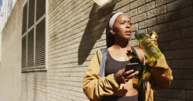 Afrika kökenli Amerikalı kadın dışarıda egzersiz yapıyor, su içiyor ve şehirde akıllı telefon kullanıyor. sağlıklı açık hava yaşam tarzı fitness eğitimi.