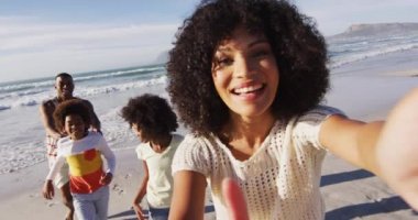 Gülümseyen Afrikalı Amerikalı aileler ve çocukları sahilde akıllı telefonlarıyla selfie çekiyorlar. Aile deniz kenarında boş vakit geçirir..