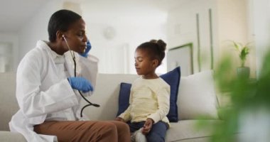 Afro-Amerikalı kadın doktor evde hasta kızla konuşuyor, steteskop kullanıyor ve gülümsüyor. Tıbbi uzman hasta ev ziyareti yapıyor.