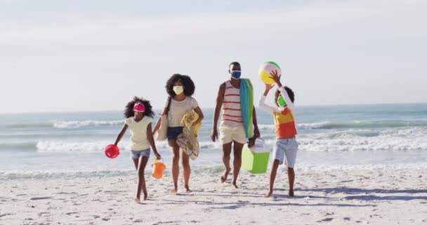 アフリカ系アメリカ人の両親と彼らの子供たちはビーチでビーチ機器を運ぶフェイスマスクを着ています コロナウイルスの時の海による家族の屋外レジャー時間Covid 19パンデミック — ストック動画