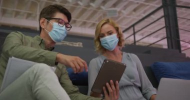 Yüz maskesi takan Asyalı bir adam ve beyaz bir kadın ofiste dijital tablet üzerinde tartışıyorlar. Coronavirus covid-19 salgını sırasında işyerinde hijyen ve sosyal mesafeler.