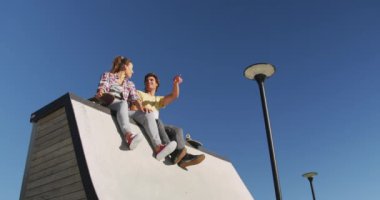 Kafkasyalı mutlu kadın ve erkek arkadaşları yüz maskesi takıyor, oturuyor ve uzaktan bir şey doğrultuyorlar. Yazın kaykay parkında takılıyorum..