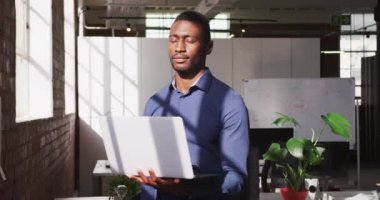 Elinde dizüstü bilgisayar olan Afrikalı Amerikalı iş adamı kameraya bakıyor ve gülümsüyor. Modern bir ofiste iş..