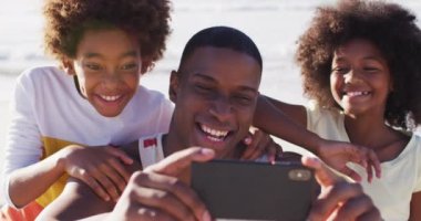 Afrikalı Amerikalı baba ve çocukları sahilde akıllı telefonlarıyla selfie çekiyorlar. Aile deniz kenarında boş vakit geçirir..