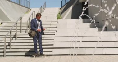 Kafkasyalı iş adamı çeşmenin arkasında kaykay yapıyor ve güneşli bir günde akıllı telefon kullanıyor. Yazın kaykay parkında takılıyorum..