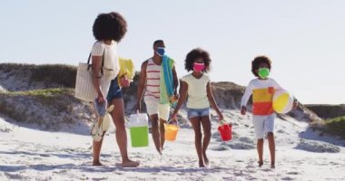 Afrika kökenli Amerikalı aileler ve çocukları plajda plaj ekipmanları taşıyan yüz maskeleri takıyorlar. Aile deniz kenarında boş vakitlerinde Coronavirus covid 19 salgını sırasında.