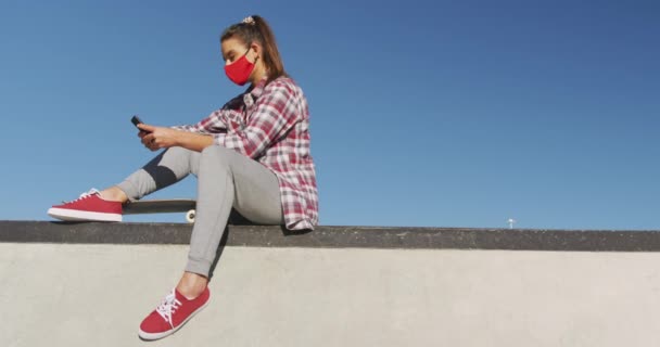 顔のマスクをした白人女性が座ってスマートフォンを使っている コロナウイルスが流行した19年の夏にスケートパークで過ごし — ストック動画