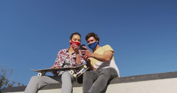 白人女性と男性の友人の顔マスクを着て 座って スマートフォンを使用しています コロナウイルスが流行した19年の夏にスケートパークで過ごし — ストック動画