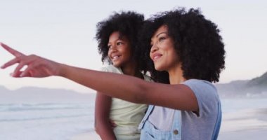 Afrika kökenli Amerikalı anne ve kızı kumsalda birbirlerine bakıp gülümsüyorlar. Aile seyahati tatil eğlence konsepti