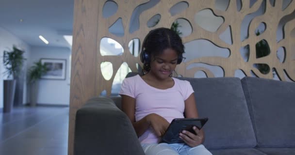 アフリカ系アメリカ人の少女は 病院のソファに座りながら デジタルタブレットを使ってヘッドフォンをしている コロナウイルスの医療は19のパンデミックの概念を覆し — ストック動画