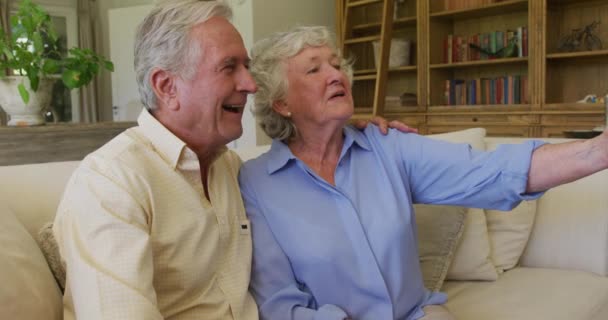 笑顔の白人のシニアカップルは自宅のソファに座ってスマートフォンから自撮りを取ります 定年退職したシニアカップルのライフスタイルのコンセプト — ストック動画