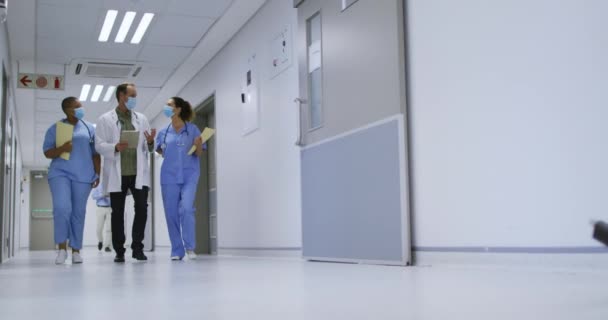 病院の廊下で話して歩く顔のマスクを身に着けている男性と女性の医師の多様なグループ コロナウイルスにおける医療 医療サービス Covid 19パンデミック — ストック動画
