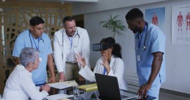 Masadaki Asyalı kadın doktor, etrafında bir grup meslektaşın toplandığı VR kulaklığı kullanıyor. ilaç, sağlık ve sağlık hizmetleri.
