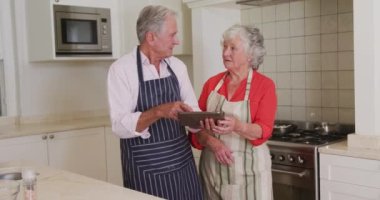 Mutfakta, yemek hazırlamadan önce önlük giymiş, tablet kullanan mutlu, beyaz bir çift. Karantina süresince evde tecrit altında..