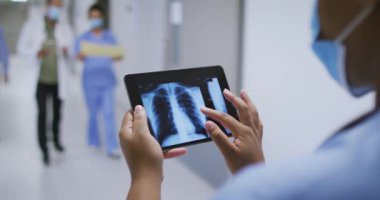 Yüzü maskeli melez bir doktor hastane koridorunda yürürken tabletteki röntgene bakıyor. Coronavirus covid 19 salgını sırasında ilaç, sağlık ve sağlık hizmetleri.