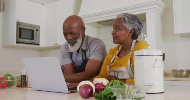アフリカ系アメリカ人のシニアカップルは 自宅のキッチンで一緒にラップトップを使用してエプロンを着ています 定年退職したシニアカップルのライフスタイルのコンセプト — ストック動画