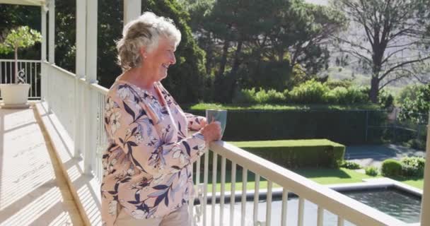 自宅のバルコニーに立ちながら笑顔でコーヒーカップを持っている白人のシニア女性 定年退職者のライフスタイルコンセプト — ストック動画