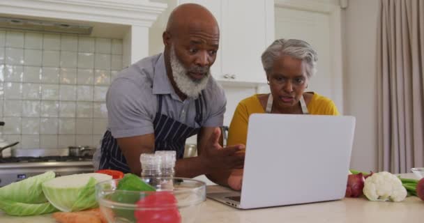アフリカ系アメリカ人の高齢者のカップルは 自宅のキッチンでラップトップでビデオ通話をしているエプロンを着ています 定年退職したシニアカップルのライフスタイルのコンセプト — ストック動画