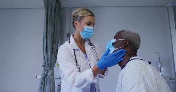 アフリカ系アメリカ人の高齢男性患者の喉を調べる顔マスクを身に着けている白人女性医師 コロナウイルスの医療は19のパンデミックの概念を覆し — ストック動画