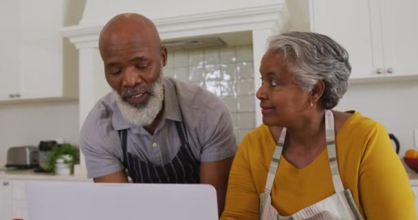 アフリカ系アメリカ人の高齢者のカップルは 自宅のキッチンでラップトップでビデオ通話をしているエプロンを着ています 定年退職したシニアカップルのライフスタイルのコンセプト — ストック動画