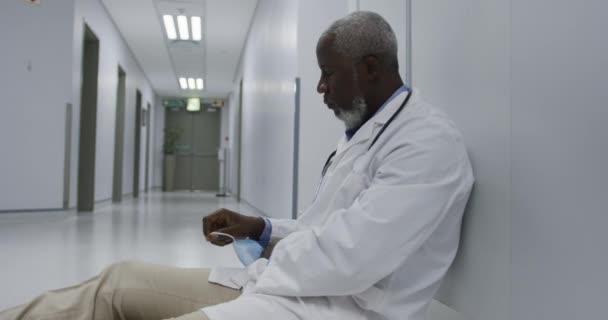 疲惫的非洲裔美国男医生坐在医院走廊上 戴着面具 Coronavirus Covid大流行病期间的医疗 保健和保健服务 — 图库视频影像