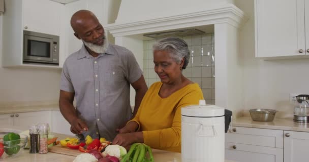 自宅のキッチンで野菜を切りながら妻を抱きしめるアフリカ系アメリカ人の先輩 定年退職したシニアカップルのライフスタイルのコンセプト — ストック動画