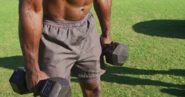 Yorgun, üstsüz Afrikalı Amerikalı adam dambılları düşürüyor ve dışarıda egzersiz yaptıktan sonra iyileşiyor. Spor sahasında spor eğitimi için çapraz eğitim.