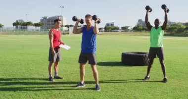 İki sağlıklı erkek ve erkek antrenörden oluşan çeşitli bir grup dışarıda egzersiz yapıyor, çömeliyor ve dambılları kaldırıyor. Spor sahasında spor eğitimi için çapraz eğitim.