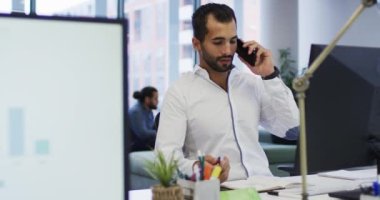 Karışık ırk işadamı masada bilgisayarla oturup meşgul bir ofiste akıllı telefondan konuşuyor. Modern bir ofiste bağımsız yaratıcı iş.