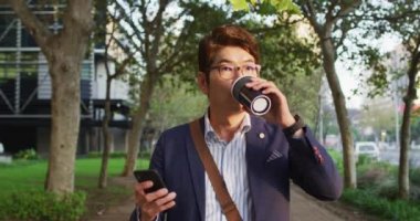 Asyalı adam parkta yürürken akıllı telefon kullanarak kahve fincanı tutuyor. iş ve yaşam tarzı kavramı