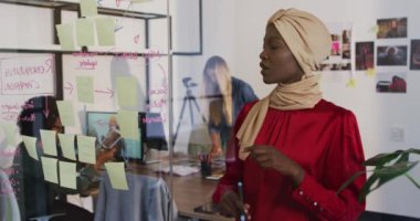 Afrikalı Amerikalı iş kadını iş arkadaşlarıyla konuşuyor ve ofisindeki cam duvara notlar yapıştırıyor. Bağımsız bir yaratıcı şirkette çalışmak.