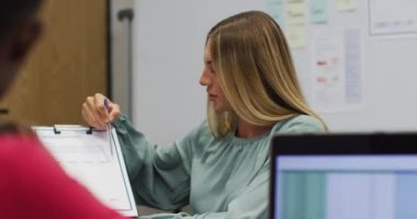 Kafkasyalı iş kadını elinde evrak işleriyle ofisteki meslektaşıyla konuşuyor ve gülümsüyor. Bağımsız bir yaratıcı şirkette çalışmak.