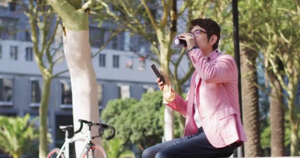 Asiatischer Mann Trinkt Kaffee Und Benutzt Smartphone Während Park Sitzt — Stockvideo