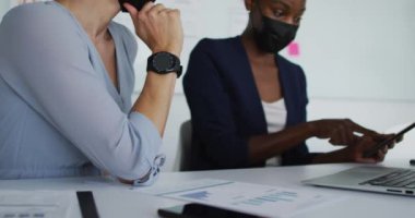 Maskeli, maske takan, masada oturmuş konuşan iki farklı bayan arkadaş. Coronavirus covid 19 salgını sırasında modern bir ofiste bağımsız yaratıcı işler..