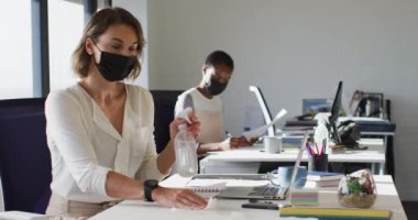Yüz maskesi takan iki farklı bayan meslektaşı ofiste masaları dezenfekte ediyor. Coronavirus covid 19 salgını sırasında modern bir ofiste bağımsız yaratıcı işler..