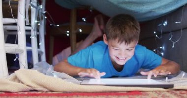 Beyaz çocuk evdeki battaniye kalesinin altında dijital tablet kullanırken gülümsüyor. aile, sevgi ve birliktelik kavramı