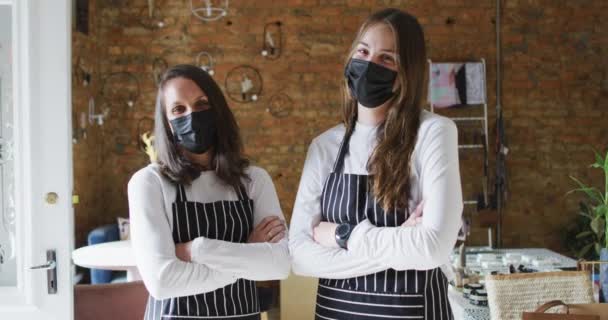 顔のマスクとエプロンを着た2人の白人女性がカメラを見ている コロナウイルスの流行期の小さな独立系カフェビジネスは — ストック動画