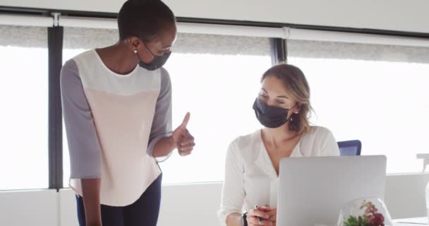 顔のマスクをした2人の多様な女性同僚がノートパソコンを見てオフィスで議論している コロナウイルスの流行期に近代的なオフィスで独立した創造的なビジネスは — ストック動画