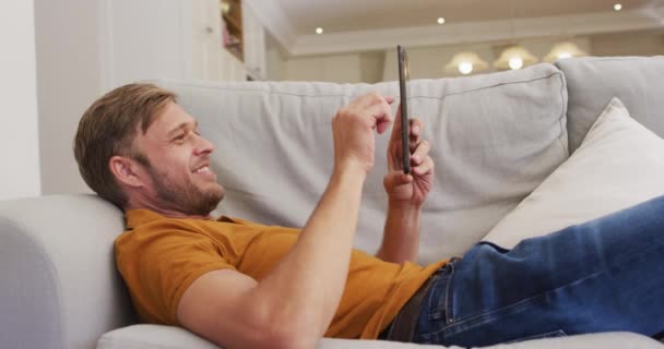 幸せな白人男性のタブレットを使用してソファの上に横たわるの肖像画 隔離されたまま家にいると — ストック動画