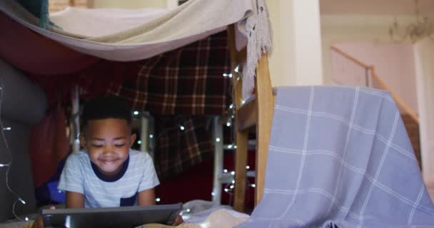 快乐的非洲裔美国男孩躺在由毛毯制成的房子里 用平板电脑 隔离检疫期间单独呆在家里 — 图库视频影像