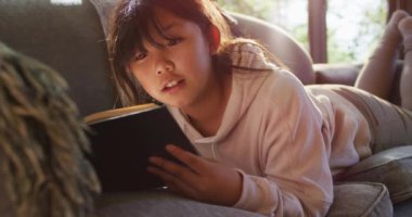 Asyalı kız evde kanepede uzanmış kitap okurken gülümsüyor. çocukluk, hobi ve boş zaman aktivite kavramı