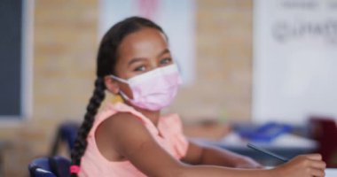 Yüz maskesi takmış, sınıfta oturmuş kameraya bakan melez bir kız çocuğunun portresi. Coronavirus covid 19 salgını sırasında ilkokuldaki çocuklar.