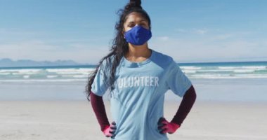 Gönüllü tişört ve yüz maskesi takıp kameraya bakan melez bir kadının portresi. Çevre koruma gönüllüleri, Coronavirus covid 19 salgını sırasında plaj temizliği..