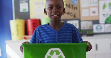 Gülümseyen Afrikalı Amerikalı okul çocuğu, elinde geri dönüşüm kutusu, sınıfta duruyor. İlkokuldaki çocuklar yazın.