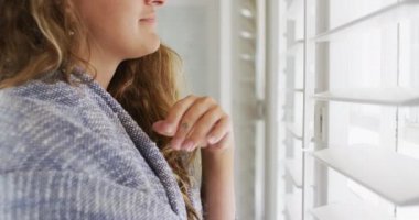 Omuzlarının üzerinde battaniyesi olan mutlu, beyaz bir kadın pencereden dışarı bakıyor ve güneşli bir evde gülümsüyor. Kırsal kesimlerin dışında basit bir evde yaşamak.