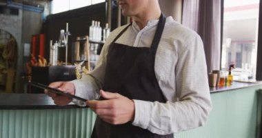Barda çalışan, tablet kullanan ve kameraya gülümseyen önlüklü beyaz adamın portresi. Bağımsız bir bar işletmesinde çalışmak, pandemik tecritten sonra tekrar açılmak.