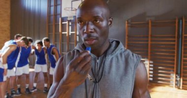 Afrika kökenli Amerikalı erkek basketbol koçu arka planda ekibiyle ıslık çalıyor. Basketbol, kapalı sahada spor eğitimi..